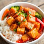 A vibrant bowl of vegetarian orange chicken featuring crispy tofu cubes, drenched in a glossy orange sauce, garnished with sesame seeds and chopped green onions, surrounded by colorful bell peppers and broccoli, served over fluffy white rice with a hint of steam rising, set against a rustic wooden table backdrop.